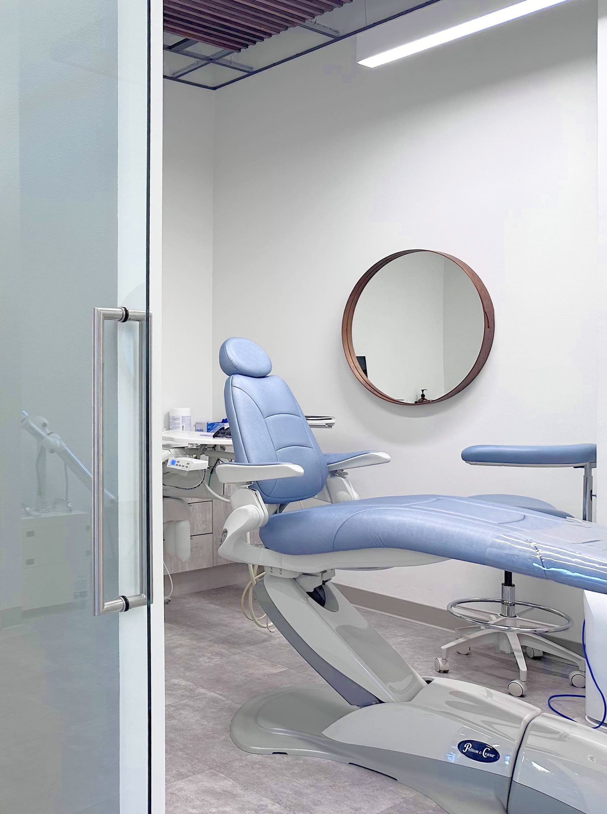 A treatment room at the San Francisco studio, with the powder blue dental chair, a round walnut mirror, and the frosted glass door